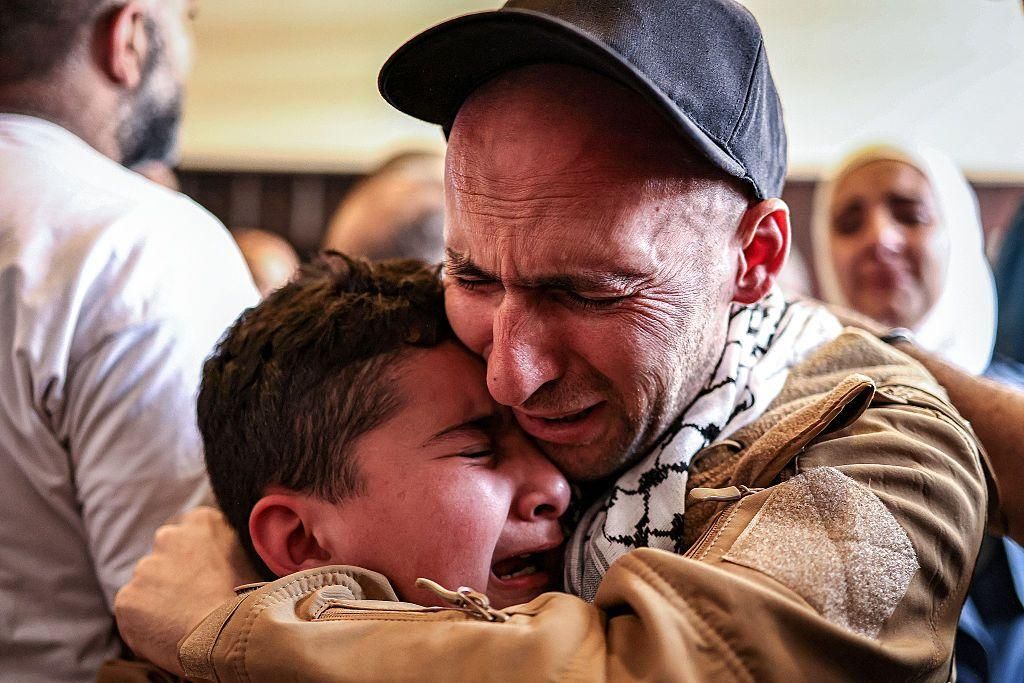 يحتضن فلسطيني يرتدي سترة بنية اللون ويلتحف بالكوفية الفلسطينية طفله، ويبكي كلاهما. في خلفية الصورة عدد من الأشخاص الآخرين.