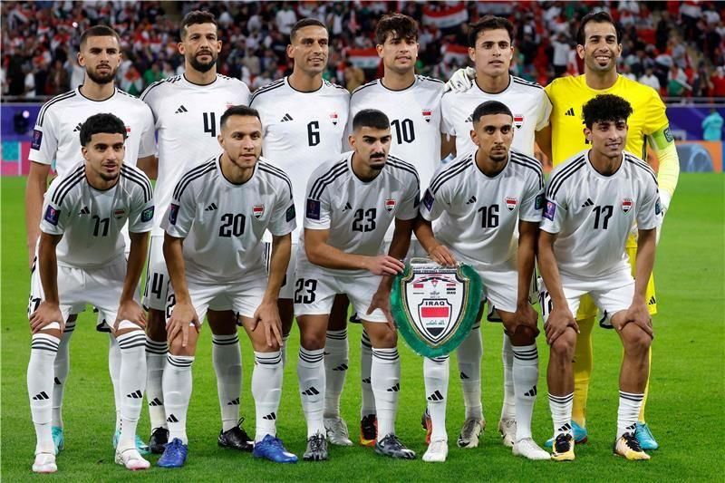 المنتخب العراقي لكرة القدم بلباسه الأبيض يحمل درعاً ويقف على أرض الملعب مستعداً لالتقاط صورة.