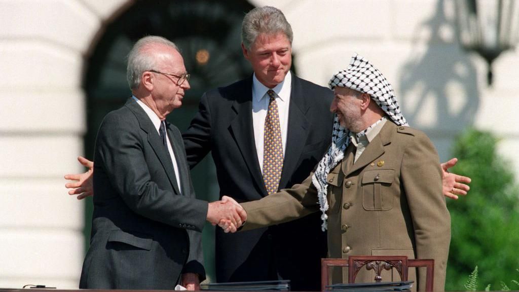 رئيس السلطة الفلسطينية ياسر عرفات (يمين) يصافح رئيس الوزراء الإسرائيلي إسحق رابين (يسار)، ويقف بينهما الرئيس الأمريكي بيل كلينتون فاتحاً ذراعيه
