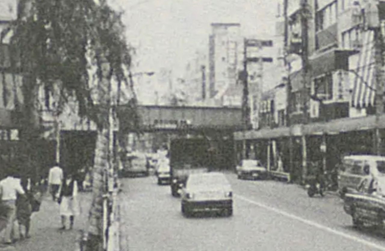 جسر سكة القطار المعلقة كما تتم مشاهدتها من الحي التجاري أمام محطة شين-أوكوبو حوالي عام 1965. المصدر: كتاب ”المدينة التجارية شينجوكو“، أرشيف مكتبة البرلمان الوطنية.